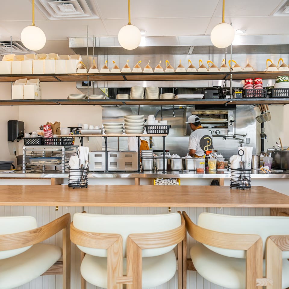 Bright restaurant counter with stacked plates, takeout boxes, hanging lights, kitchen equipment, and a worker preparing food.