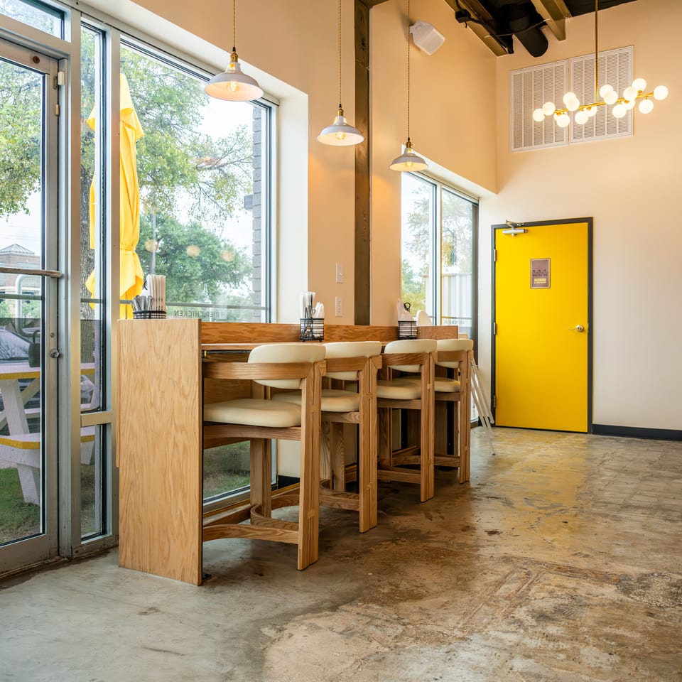 Bright restaurant interior with wooden tables and chairs, large windows, pendant lights, and a vivid yellow door.