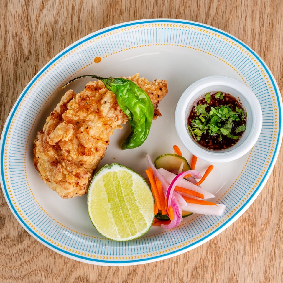 Crispy fried chicken served with a lime half, green pepper, pickled vegetables, and herb-topped dipping sauce.