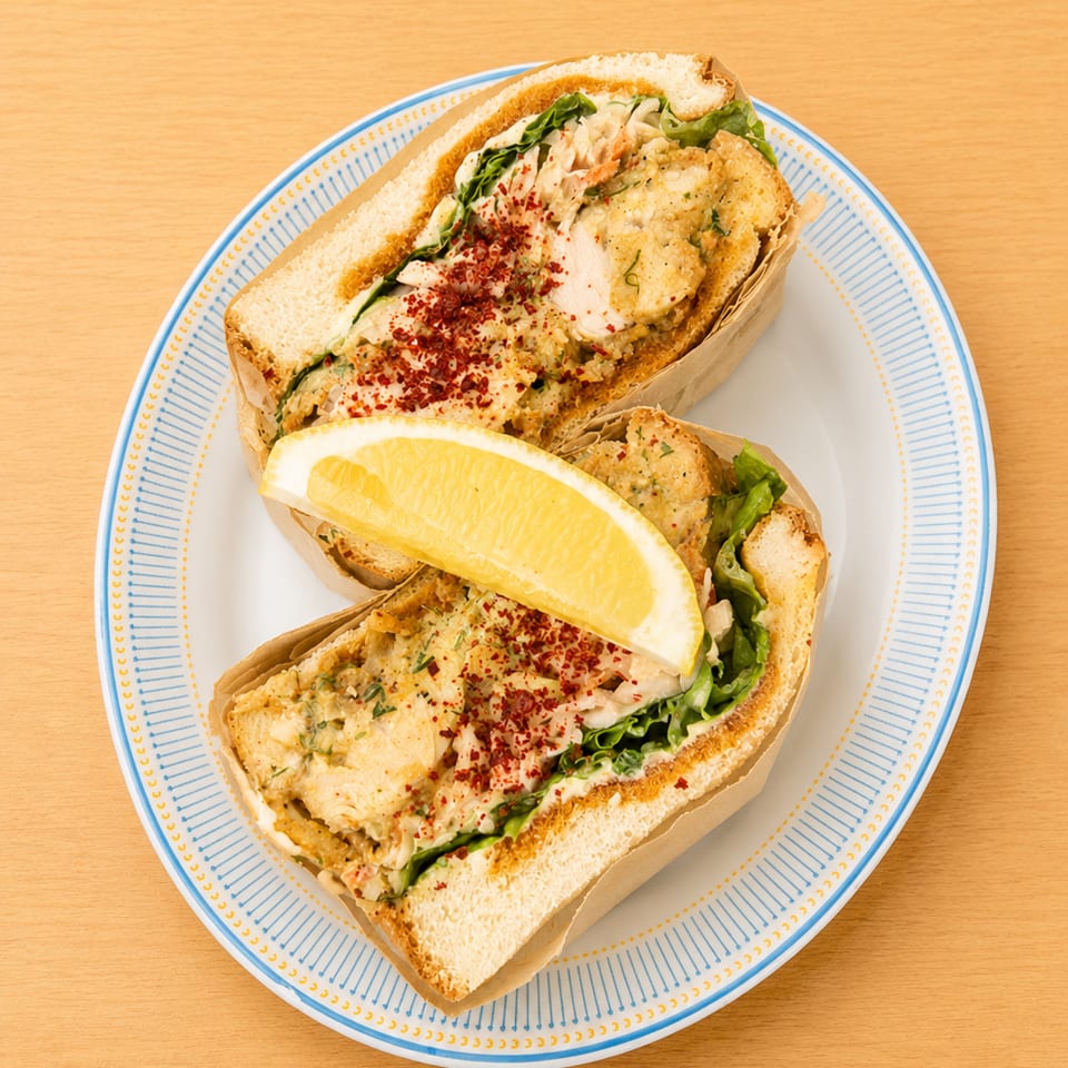 Two halves of a fried fish sandwich with lettuce, red spice, and a lemon wedge on a plate.