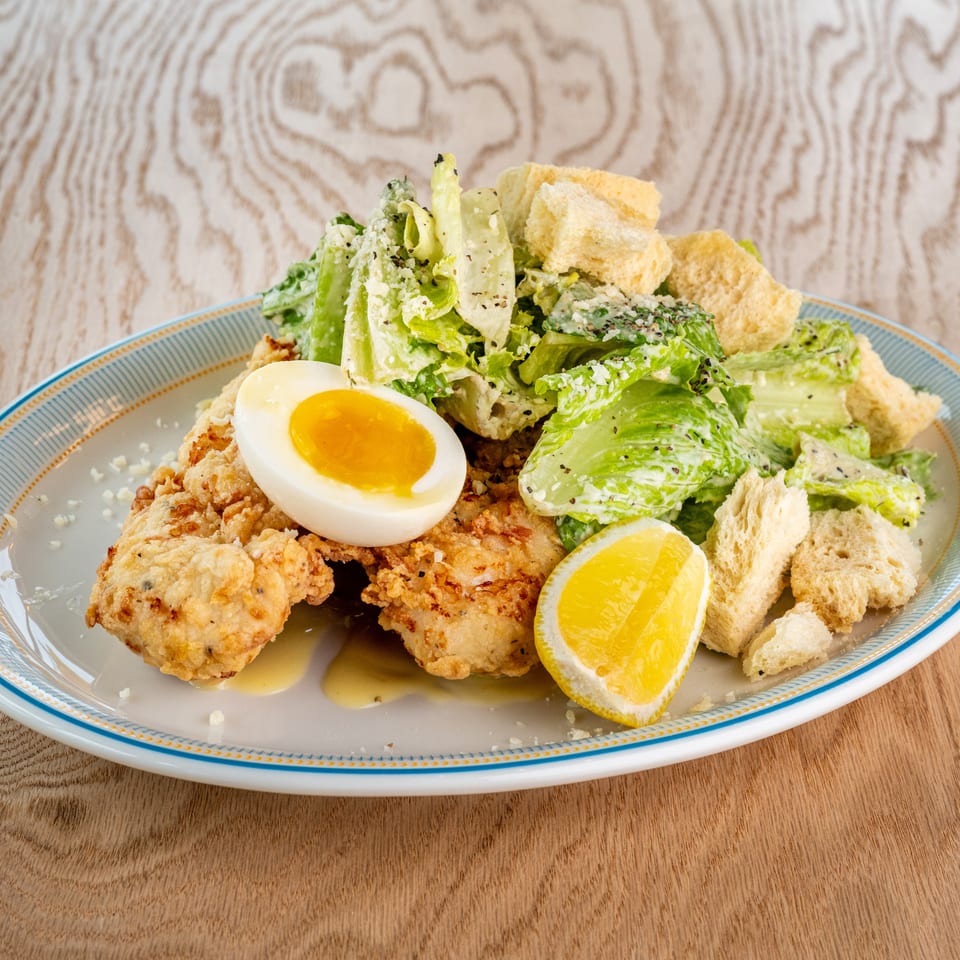 Plate of crispy fried chicken topped with a soft-boiled egg, romaine salad, croutons, and lemon wedge.