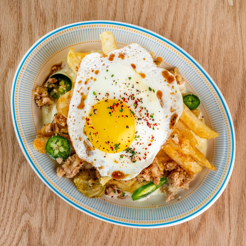 Overhead view of crispy fries topped with a fried egg, crumbled meat, jalapeños, herbs, spices, and sauce on a patterned plate.