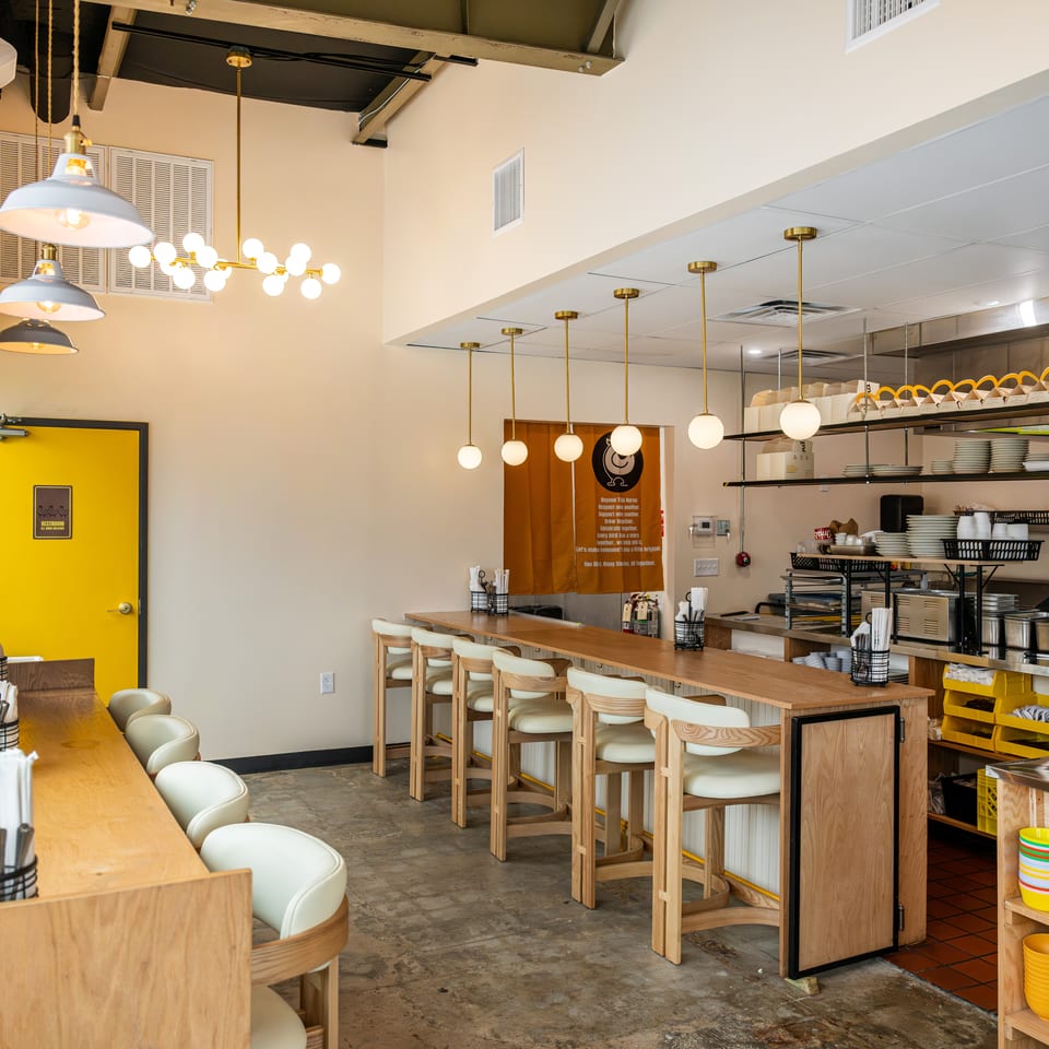Bright modern restaurant interior with wooden counter seating, pendant lights, open kitchen shelves, and colorful dishes.