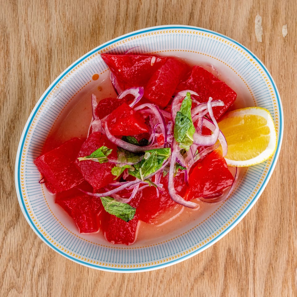 Plate of watermelon salad topped with red onion, mint, and a lemon wedge on a wooden table.