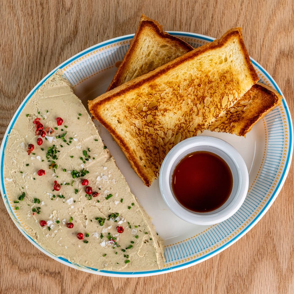 Overhead view of toasted bread, creamy pâté garnished with herbs and peppers, and a cup of dark dipping sauce on a patterned plate.