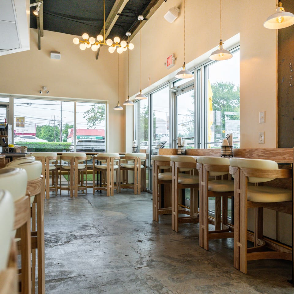 Bright restaurant interior with wooden tables, upholstered chairs, pendant lights, large windows, and a beverage station.