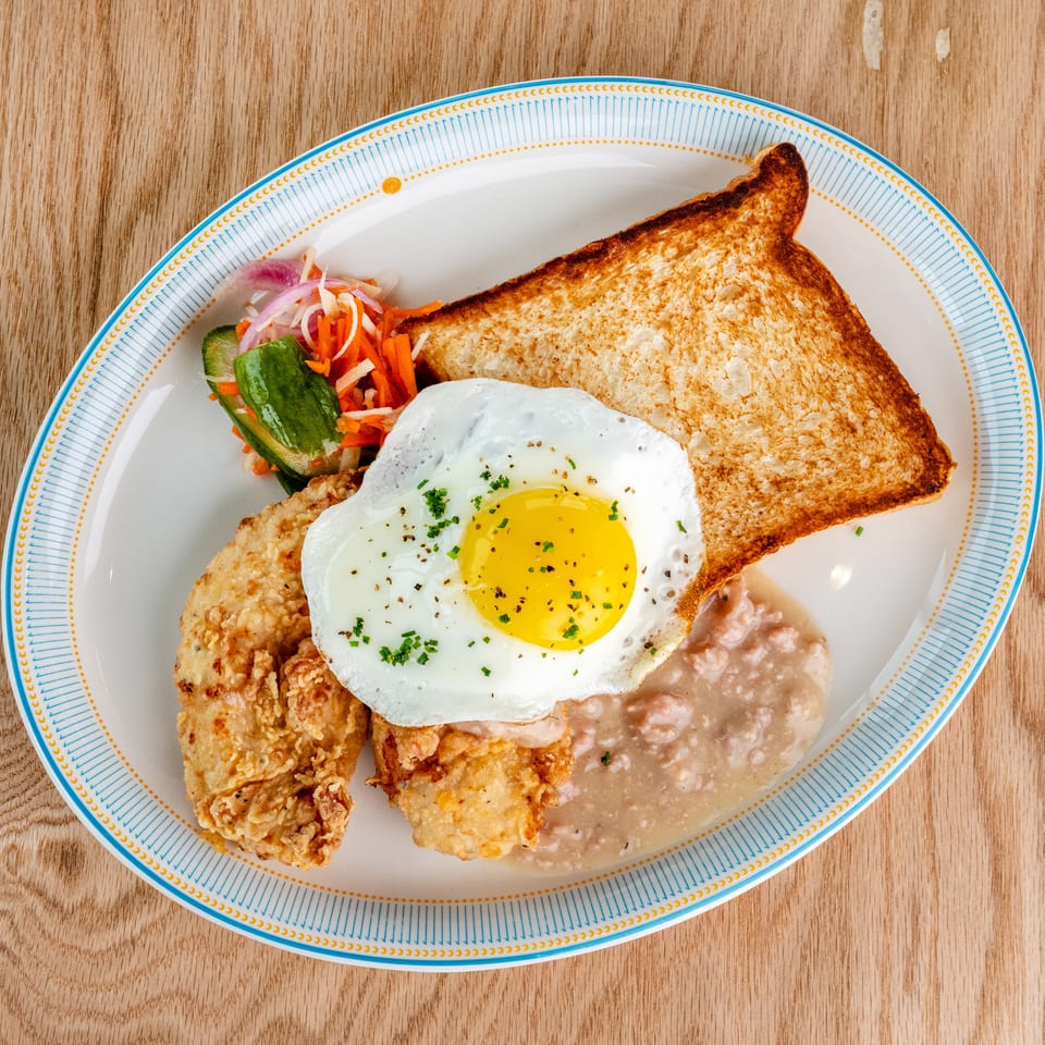 Crispy fried chicken topped with a sunny-side-up egg, toast, refried beans, and pickled vegetables on a patterned plate.