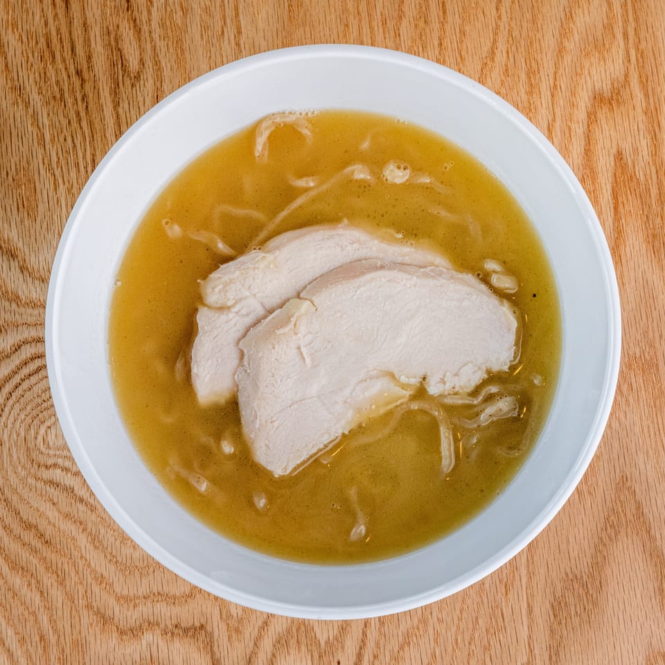 White bowl of golden broth with sliced chicken and noodles on a wooden table.