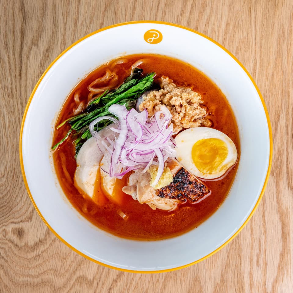 Top-down view of spicy noodle soup topped with chicken, minced meat, greens, red onion, tofu, and a halved boiled egg.