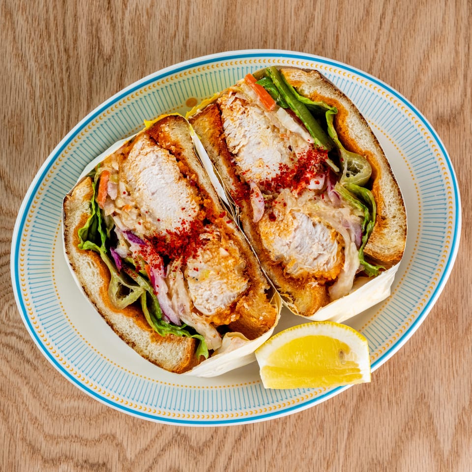 Two toasted chicken sandwiches with lettuce, vegetables, creamy sauce, and red pepper flakes on a patterned plate with lemon wedges.