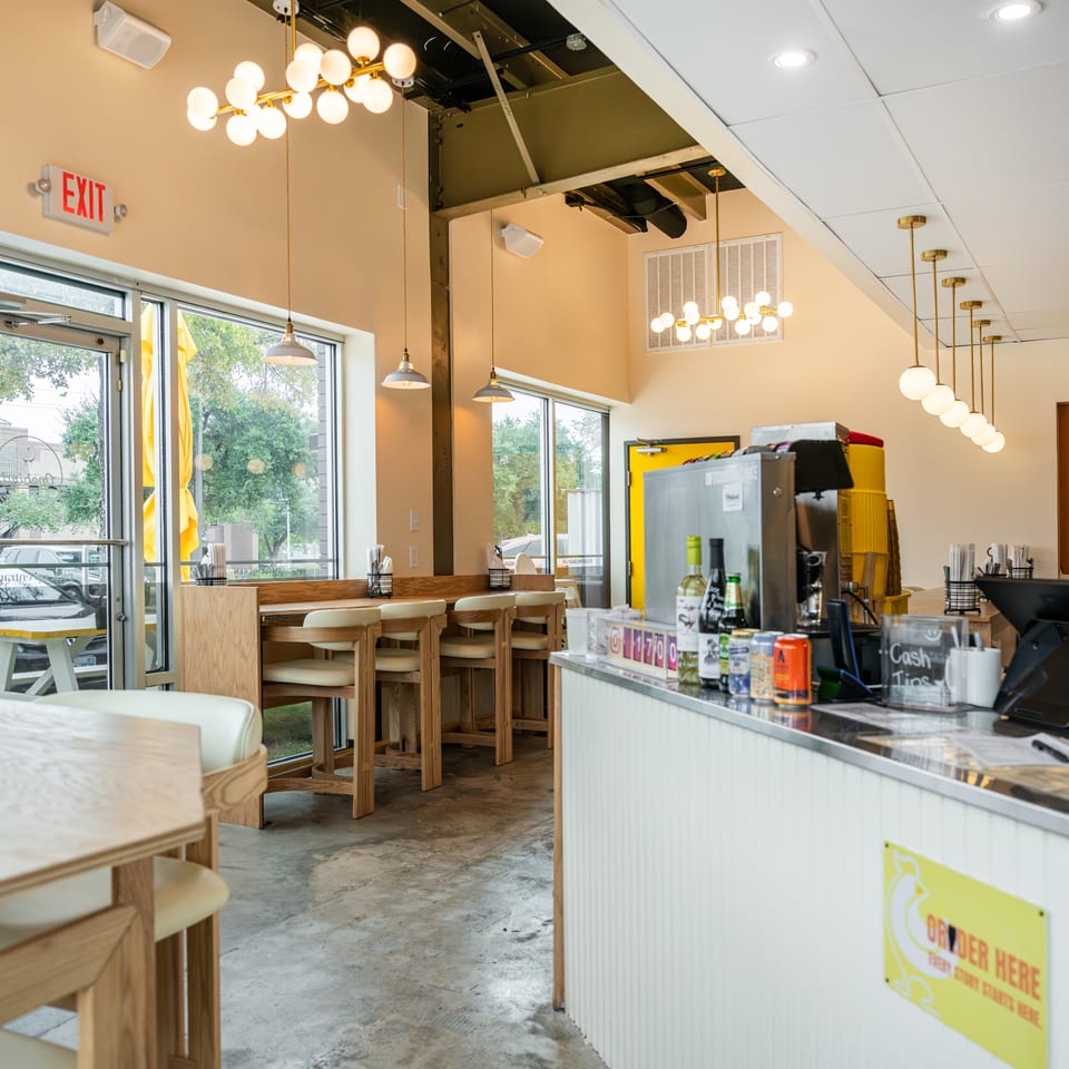 Bright restaurant interior with wooden seating, globe pendant lights, large windows, and a counter displaying drinks and ordering signs.