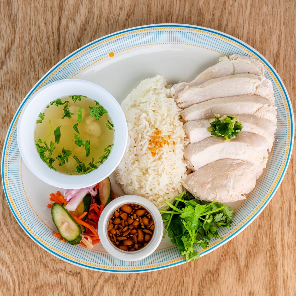 Plate of sliced poached chicken, white rice, herb broth, dipping sauce, pickled vegetables, and fresh cilantro.
