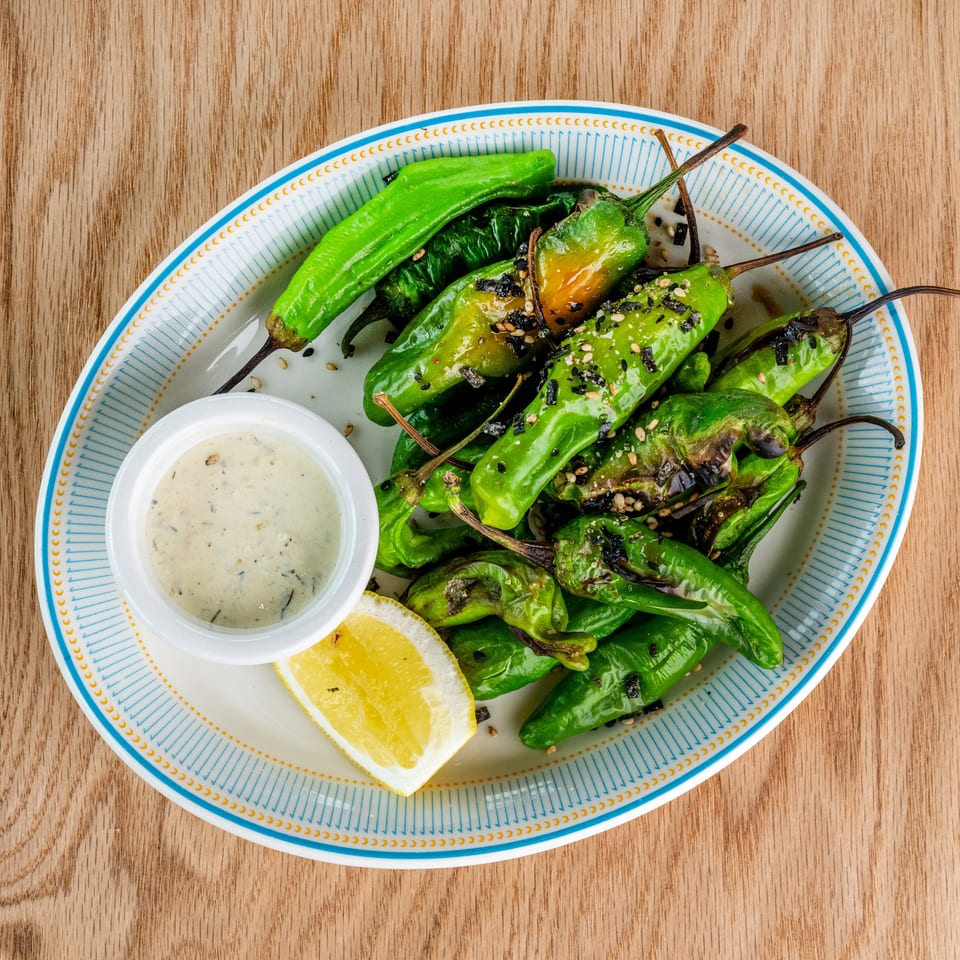 Grilled green peppers topped with sesame seeds and seaweed, served with creamy dipping sauce and lemon wedges on a patterned plate.