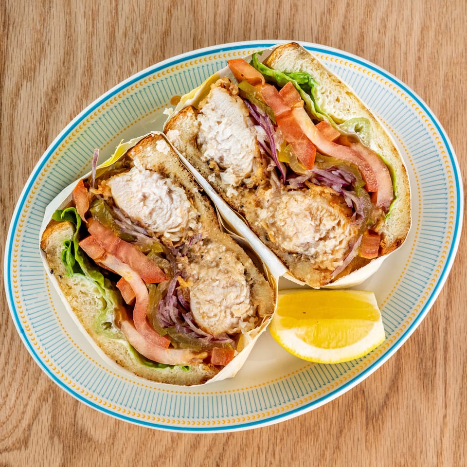 Two fried chicken sandwiches with lettuce, tomato, pickles, onions, and sauce served with lemon wedges on a patterned plate.