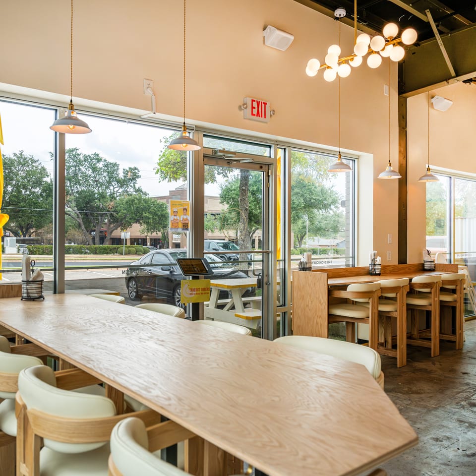 Bright modern restaurant interior with wooden tables, cream chairs, pendant lights, large windows, and yellow accents.