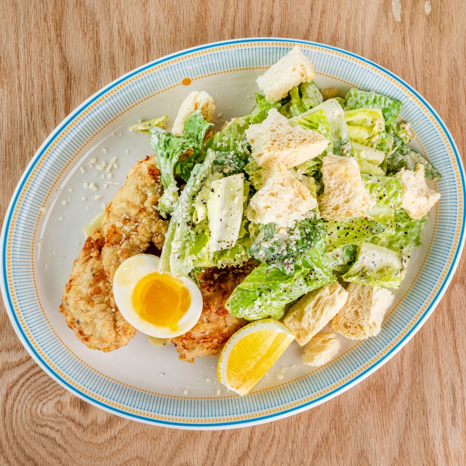 Overhead view of a breaded chicken cutlet with Caesar salad, croutons, soft-boiled egg, and lemon on a patterned plate.