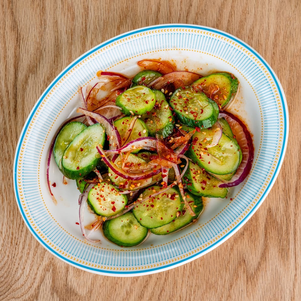 Cucumber and red onion salad seasoned with chili flakes on a patterned plate atop a wooden table.