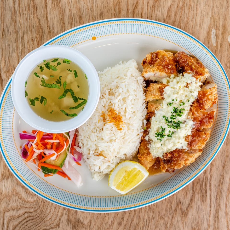 Plate of crispy fried chicken with creamy sauce, white rice, pickled vegetables, lemon, and herb-garnished broth on a wooden table.